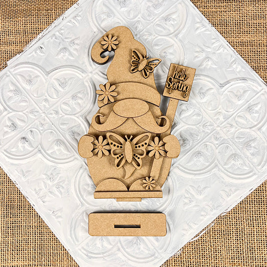 The Spring Butterfly Gnome Shelf Sitter from Janet's Craft Corner is a DIY craft kit featuring a gnome holding a "Hello Spring" sign. Ideal for home décor, it includes floral and butterfly accents on a textured white background with a base slot.