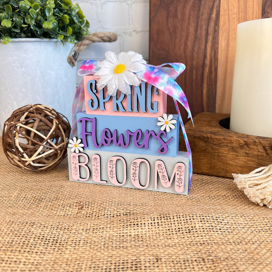 The Spring Word Stack from Janet's Craft Corner captures the essence of spring with pink and purple lettering spelling out "Spring Flowers Bloom." Adorned with a white daisy and colorful ribbon, it is beautifully complemented by a woven ball, candle, and nearby potted plant to create an enchanting seasonal display.