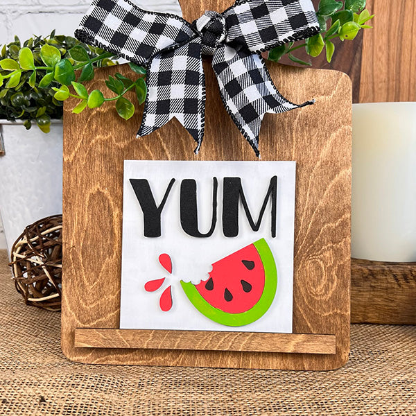 Janet's Craft Corner Breadboard Swappable Squares Holder DIY Craft Kit displays a "YUM" sign above a bitten watermelon. Featuring a black-and-white checkered bow and green leaves, it’s perfect for your interchangeable square signs décor.
