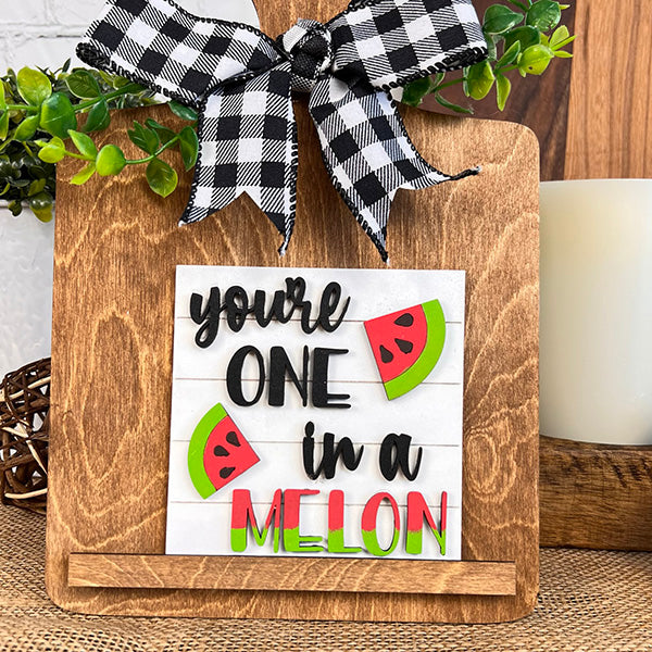 The Breadboard Swappable Squares Holder DIY Craft Kit by Janet's Craft Corner features "You’re one in a melon" with watermelon art, plaid bow, and greenery. This charming décor rests on wood next to a candle.