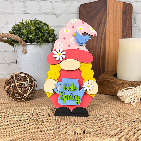 The "Hello Spring Girl Gnome" from Janet's Craft Corner is a charming addition to your spring decor. This colorful figurine features yellow braids, a pink hat adorned with daisies, and holds a "Hello Spring" sign. A blue bird sits perched on her hat, while nearby are placed rustic wooden decorations and a plant in a white pot.