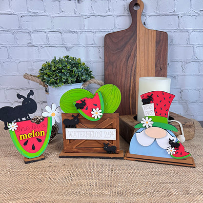 Janet's Craft Corner presents a charming decorative display featuring farmhouse decor with wooden summer-themed items: a watermelon slice engraved with "one in a melon," ants atop leaves in a crate marked "watermelon 25¢," and a gnome figurine sporting a strawberry hat. A plant and DIY kit complement the scene, alongside Watermelon Shelf Sitters standing in the background.