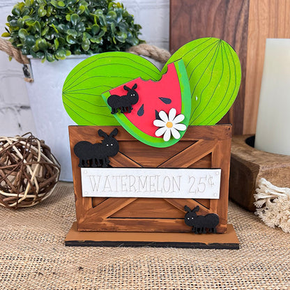 This charming decoration from Janet's Craft Corner, part of the Watermelon Shelf Sitters collection, showcases a wooden display featuring a watermelon slice adorned with leaves and ants beneath a "WATERMELON 25¢" sign. A lovely daisy accents the slice, complemented by a woven ball and potted plant in the background.