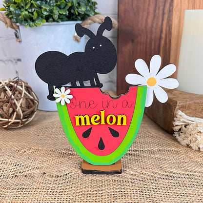 Introducing the Watermelon Shelf Sitters from Janet's Craft Corner: a delightful decorative piece featuring an ant perched on a watermelon slice with the playful phrase "One in a melon." Perfect for farmhouse decor, this unique part of an enchanting DIY kit comes with a bite-marked slice embellished with a white daisy. Included are additional accents like a small plant, wooden decor elements, and a woven ball placed atop burlap for added charm.