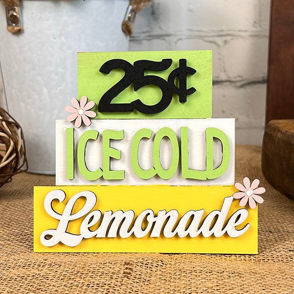 The Summer Word Stacks by Janet's Craft Corner features a decorative sign made up of three layered boards, with "25¢" in black, "Ice Cold" in green, and "Lemonade" in white. Each layer has a small pink flower accent, reminiscent of farmhouse-style decor. The backdrop includes a brick wall and woven basket to evoke rustic charm.