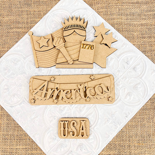 Wooden cutouts on a textured background celebrate Americana, showcasing the Statue of Liberty, an American flag with stars, a "1776" sign, the word "America" adorned with stars, and "USA" below. Perfect for Janet's Craft Corner's America Interchangeable Insert DIY home decor kit.