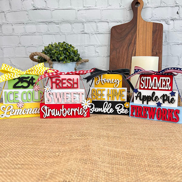 The Summer Word Stacks from Janet's Craft Corner offers charming farmhouse-style decor with phrases such as "Ice Cold Lemonade" and "Honey Bee Hive." Each DIY kit comes with colorful text and bows, all set against a burlap surface on a rustic brick wall, making it ideal for creating your own summer-themed decorative blocks.