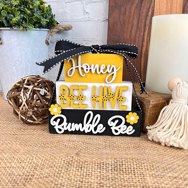 This product from Janet's Craft Corner, called the Summer Word Stacks, features a charming wooden block display inscribed with the words "Honey," "BUMBLE BEE," and "Bee Hive" in various fonts. Decorated with a black ribbon, yellow accents, and small floral designs, this piece adds rustic charm to any collection of farmhouse-style decor when placed on a burlap-covered surface.