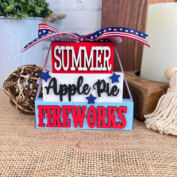 Introducing the Summer Word Stacks from Janet's Craft Corner: This decor wooden sign features a farmhouse-style with a patriotic touch. It comes in a red, white, and blue palette decorated with ribbon, displaying "Summer," "Apple Pie," and "Fireworks" in charming word stacks. Star embellishments add an inviting seasonal flair to this design.