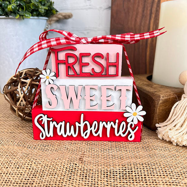 Janet's Craft Corner presents the Summer Word Stacks, featuring a decorative block stack with "Fresh Sweet Strawberries" in red and white. This charming piece is adorned with small daisies and finished with a red checkered ribbon on top. Ideal for farmhouse-style decor, it sits on a burlap surface, capturing the essence of summer with rustic charm.