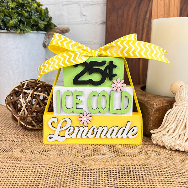 Introducing the Summer Word Stacks by Janet's Craft Corner, a decorative sign showcasing a vibrant yellow and green design with the phrase "25¢ Ice Cold Lemonade." This charming piece is accented by delicate pink flowers and an elegant yellow bow, all beautifully arranged on a burlap surface. Ideal for enhancing your farmhouse-style decor, this lovely addition brings a rustic charm to your summer setting.