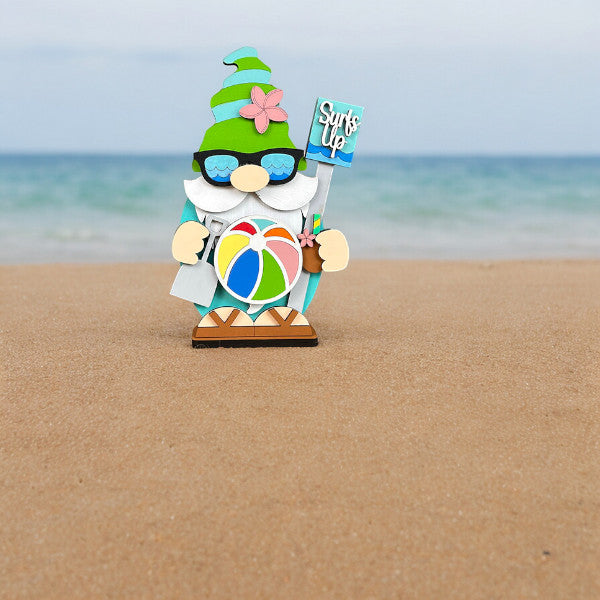 Janet's Craft Corner offers the Beach Gnome Shelf Sitter DIY home decor craft kit, featuring a whimsical gnome on a sandy beach with sunglasses, a green hat adorned with a pink flower, and holding a "Surf's Up" sign and beach ball against an ocean backdrop.