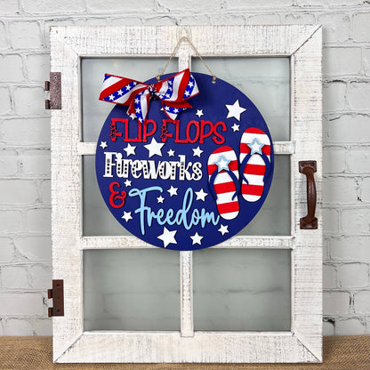The Patriotic Flip Flops Door Hanger from Janet's Craft Corner showcases a delightful design featuring a rustic white window frame with a circular blue board adorned with white stars and red and white flip flops. The piece is embellished with Americana-inspired red, white, and blue letters that read "Flip Flops, Fireworks & Freedom," adding a touch of patriotic flair to your space.