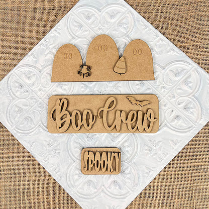 Featuring the "Boo Crew Interchangeable Insert" DIY home decor kit from Janet's Craft Corner, this hand-painted craft set includes wooden ghost shapes, a spider, and a candy corn on a textured background. It also showcases the phrases "Boo Crew" with a bat and "Spooky," making it an ideal interchangeable accessory for adding festive charm to any space.