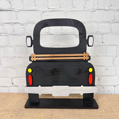 The Antique Truck Interchangeable Base - Painted by Janet's Craft Corner is a black wood craft cutout seen from behind, featuring red and yellow tail lights. It sits on burlap with a white brick wall backdrop—ideal for interchangeable decor.