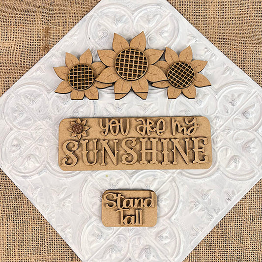 Sunflower accents on a textured backdrop: featuring three sunflowers at the top, a central plaque displaying "YOU ARE MY SUNSHINE," and a smaller plaque with "Stand Tall" positioned below. This delightful DIY kit from Janet's Craft Corner, known as the Sunflower Interchangeable Insert, is ideal for fans of versatile home decor themes.