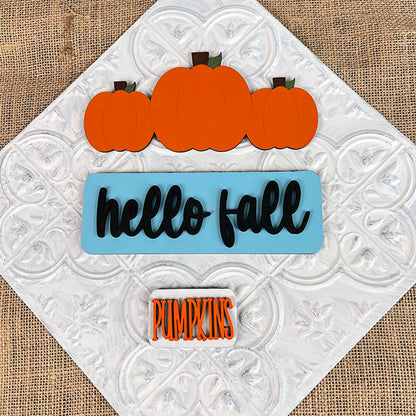 Introducing the **Hello Fall Interchangeable Insert** by *Janet's Craft Corner*: a hand-painted DIY home decor kit. This decorative sign showcases three orange pumpkins along with the phrase "hello fall" artfully scripted in black on a blue background, complemented by "PUMPKINS" elegantly displayed in orange letters. Set on a white textured burlap backdrop, this kit is an ideal addition to your seasonal decor collection.