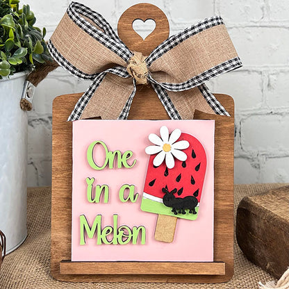 The Breadboard Swappable Squares Holder DIY Craft Kit from Janet's Craft Corner displays a pink "One in a Melon" sign with watermelon popsicle, daisy, and ant—perfect summer décor that’s compatible with swappable square signs for all seasons.