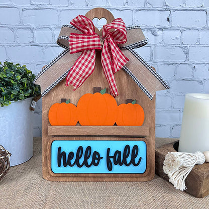 The "Hello Fall Interchangeable Insert" from Janet's Craft Corner is an ideal seasonal decoration. This DIY home decor kit includes a wooden cutting board adorned with three orange pumpkins and a charming "hello fall" sign. A red and brown plaid bow sits atop, making it perfect for placement on any table alongside a green plant, white candle, and twine ball against a brick background.