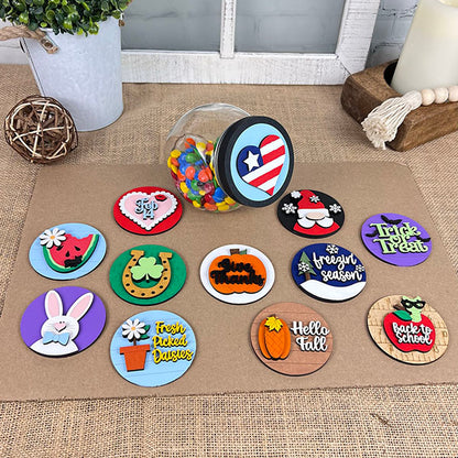 On a table, an array of vibrant, decorative magnets from Janet's Craft Corner's Candy Jar Interchangeable Set enhances the home décor with designs featuring seasonal themes such as Halloween, Christmas, Easter, and patriotic symbols. In the background, a candy jar adds to the scene with its colorful contents spilling across the surface.