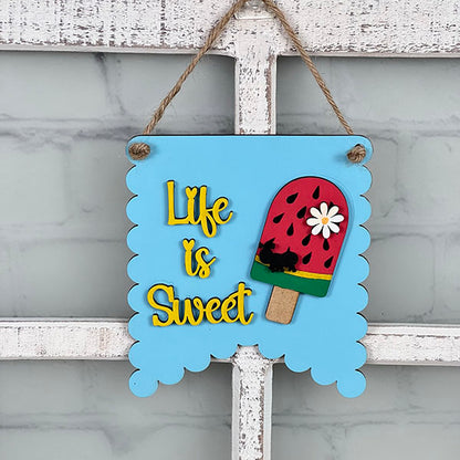 A charming addition to your home decor, a light blue scalloped-edged sign from the Summer Mini Signs collection by Janet's Craft Corner adorns a white window frame. It showcases a colorful popsicle design with floral and seed accents, accompanied by the cheerful yellow phrase "Life is Sweet.