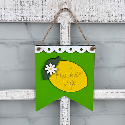 A decorative "Summer Mini Sign" from Janet's Craft Corner hangs on a rustic white door, showcasing a green banner with a vibrant yellow lemon cutout at its center. The lemon, beautifully embellished with a white flower and leaf, is inscribed with the words "Pucker Up," making it an ideal addition to your home decor or DIY kit collection.