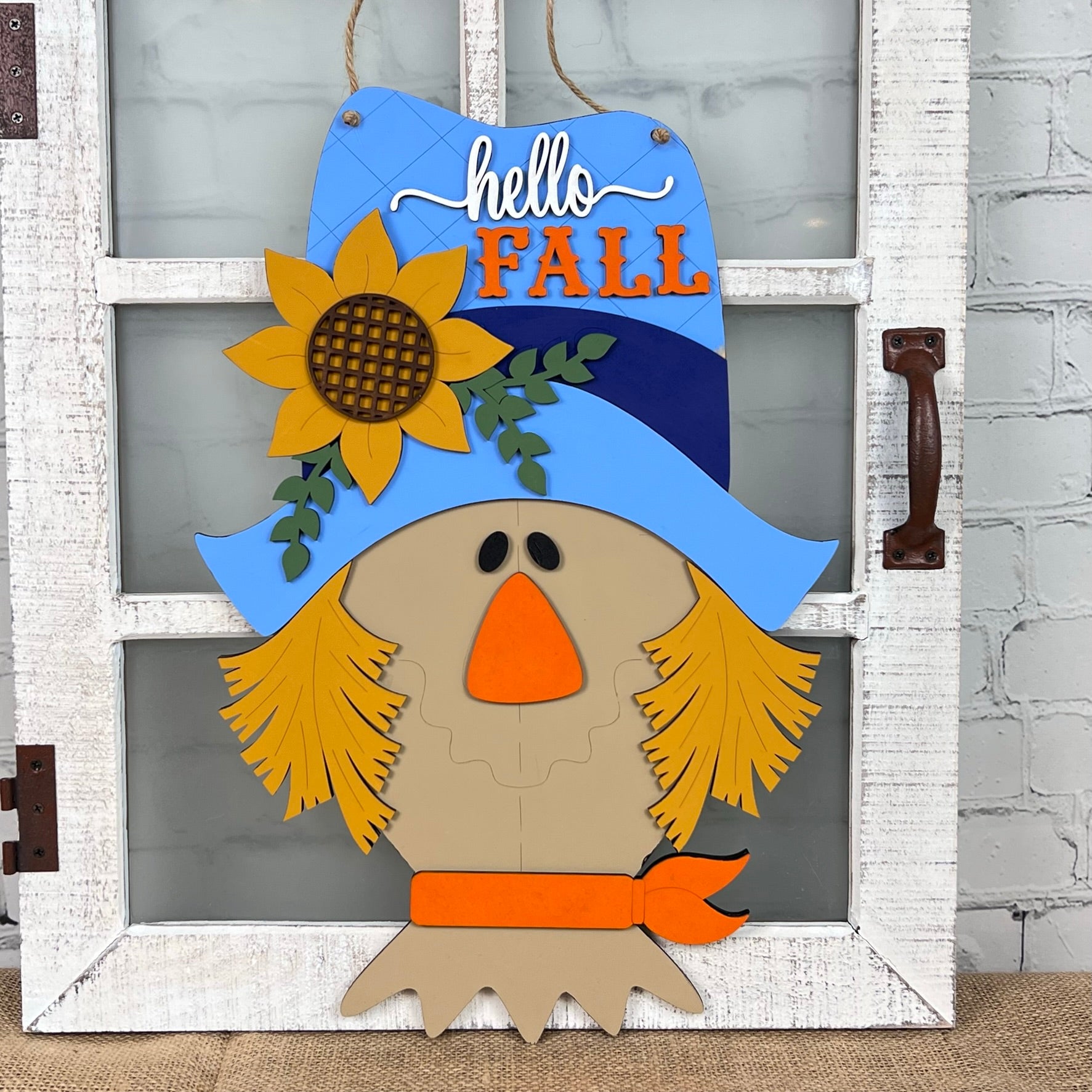 The Scarecrow Door Hanger by Janet's Craft Corner is a decorative wooden sign shaped like a scarecrow's face, designed to add charm to your door. It features a blue hat adorned with a sunflower and the phrase "hello FALL" in white and orange, all suspended on a rustic white frame against a brick background—an ideal addition to your home décor.