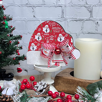 A festive scene showcases a decorated red and white Christmas card featuring Santa Claus on a stand, complete with the Chunky Santa Hat by Janet's Craft Corner. The setup is enhanced with a striped bow and bell. Surrounding it are holiday home decor items such as a candle, evergreen branches, pine cones, and red berries.