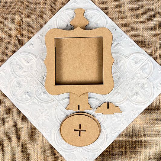 Resting on a white embossed background, Janet's Craft Corner presents the Drop In Interchangeable Squares Holder, featuring an unfinished wooden frame with ornate edges. Additional components include a circular base with a plus cutout and a small semi-circular stand, while the textured burlap backdrop adds to this DIY kit's charm.