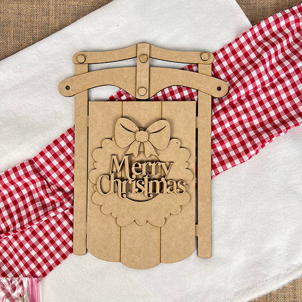 The Merry Christmas Sled DIY Craft Kit from Janet's Craft Corner adds festive charm to a white surface with its bow cutout, resting on a red and white checkered cloth and paired with a red striped accent—perfect for your holiday décor.