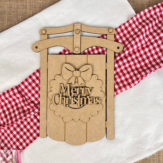 The Merry Christmas Sled DIY Craft Kit from Janet's Craft Corner adds festive charm to a white surface with its bow cutout, resting on a red and white checkered cloth and paired with a red striped accent—perfect for your holiday décor.