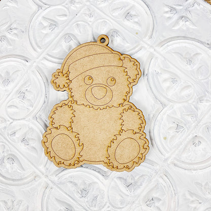 A wooden cutout of a teddy bear wearing a Santa hat rests on a white textured surface, making it an ideal choice for fans of Janet's Craft Corner's Color Your Own Ornament | DIY Decoration Kits. The charming and straightforward design includes etched details that outline its features, encouraging you to use your creativity to color the ornament and personalize your home décor.