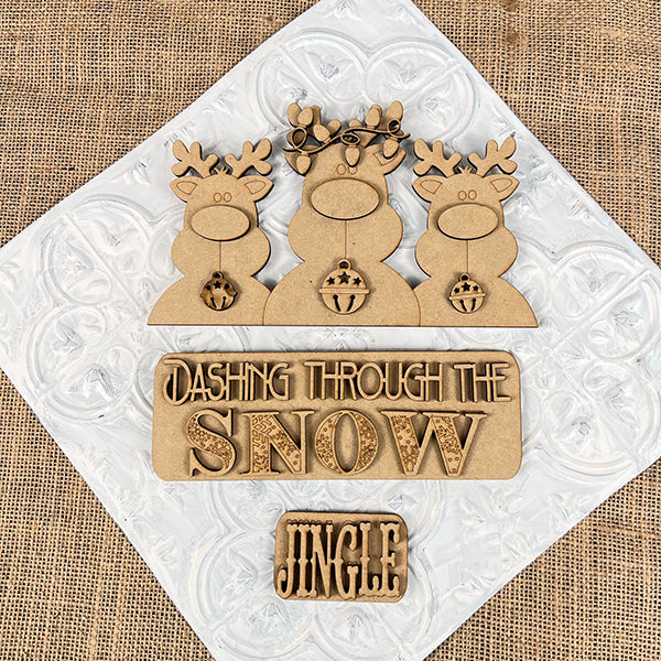 The Reindeer Interchangeable Insert from Janet's Craft Corner features three antlered reindeer set against a textured burlap background, with signs that read "Dashing Through the Snow" and "Jingle." As a DIY home decor kit, it is perfect for adding festive charm to any space.