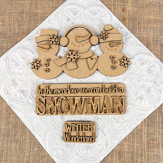 On a textured surface, the Snowman Interchangeable Insert from Janet's Craft Corner showcases wooden snowmen and text decorations with messages such as "In the meadow we can build a SNOWMAN" and "WINTER Wonderland." This hand-painted DIY home decor kit includes snowmen adorned with scarves, hats, and snowflake decorations for unique interchangeable home accents.