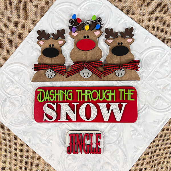 Janet's Craft Corner presents the Reindeer Interchangeable Insert DIY home decor kit, featuring three charming cartoon reindeer with red noses, plaid bow ties, antlers, and bells. This delightful piece adds a festive touch to your holiday sign with the message "Dashing Through The Snow" and "Jingle" at the bottom, making it ideal for creating a warm and inviting seasonal ambiance.