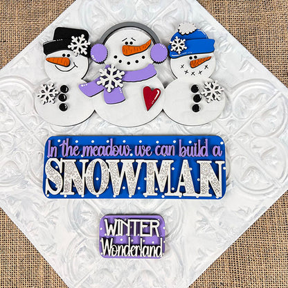 Using the Snowman Interchangeable Insert DIY home decor kit from Janet's Craft Corner, three snowmen adorned with hats and scarves stand against a decorative snowy background. A sign reading "In the meadow, we can build a SNOWMAN" is depicted in purple and blue letters, while below it, a smaller sign declares "WINTER Wonderland." This scene is beautifully accented with snowflakes for an interchangeable home decor feel.