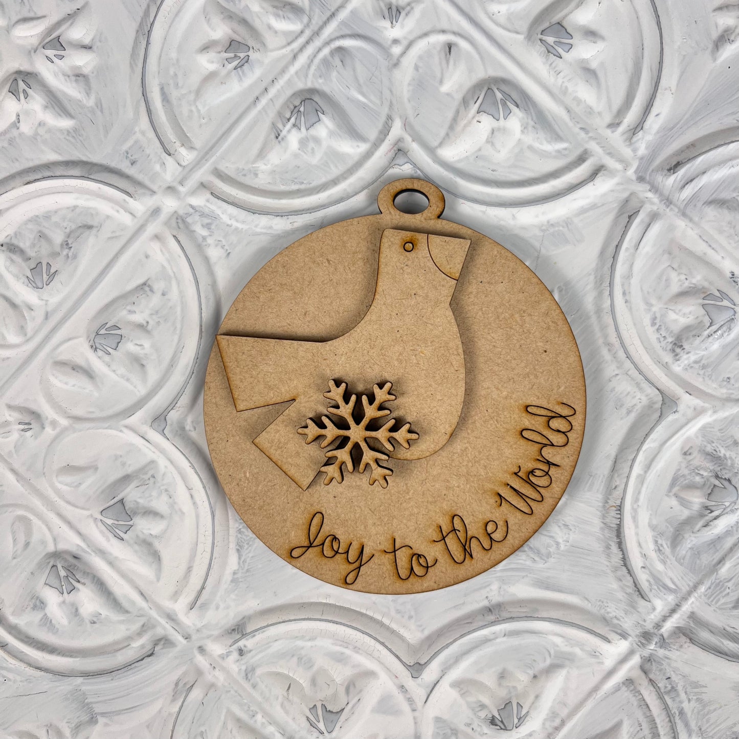 Introducing Janet's Craft Corner's Round Christmas Ornaments from the DIY Decoration Kits! This delightful wooden ornament is shaped like a bird and adorned with a snowflake, featuring "joy to the world" engraved on it. It's beautifully complemented by a textured white background, making it a charming addition to your holiday decor.