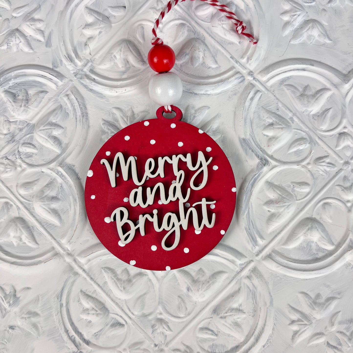 This charming piece of home décor from Janet's Craft Corner, included in the Round Christmas Ornaments | DIY Decoration Kits, features festive "Merry and Bright" text in white cursive on a red background with polka dots. It comes attached to a string adorned with red and white beads and placed against a textured white surface, making it perfect for your crafting projects!