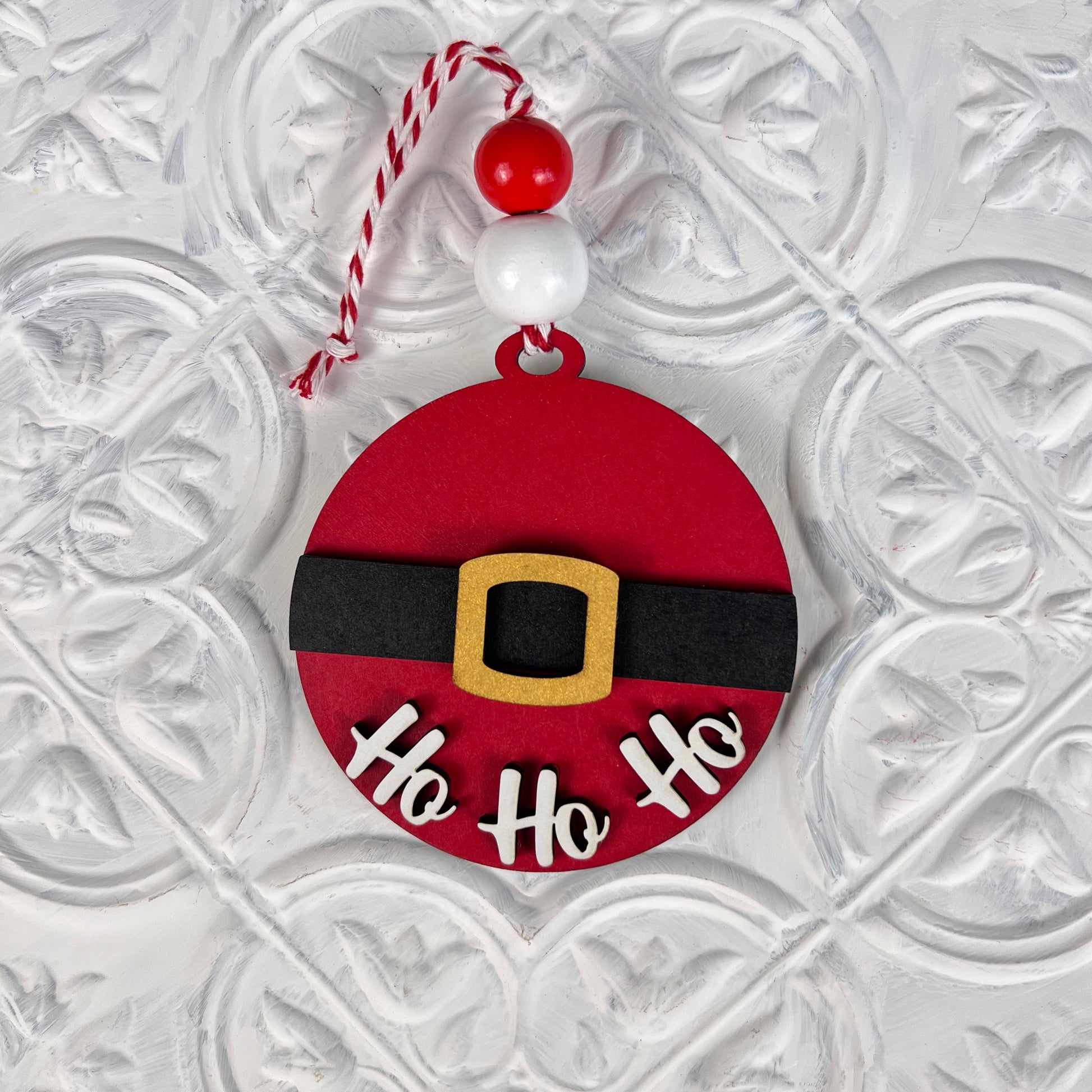 Introducing Janet's Craft Corner's Round Christmas Ornaments from the DIY Decoration Kits collection. This ornament features a vibrant red design with a black belt and gold buckle, reminiscent of Santa's iconic outfit. The phrase "Ho Ho Ho" is elegantly scripted in white below the belt, making it an ideal choice for infusing festive spirit into your home décor. It hangs gracefully from a string decorated with red and white beads, set against a textured background.