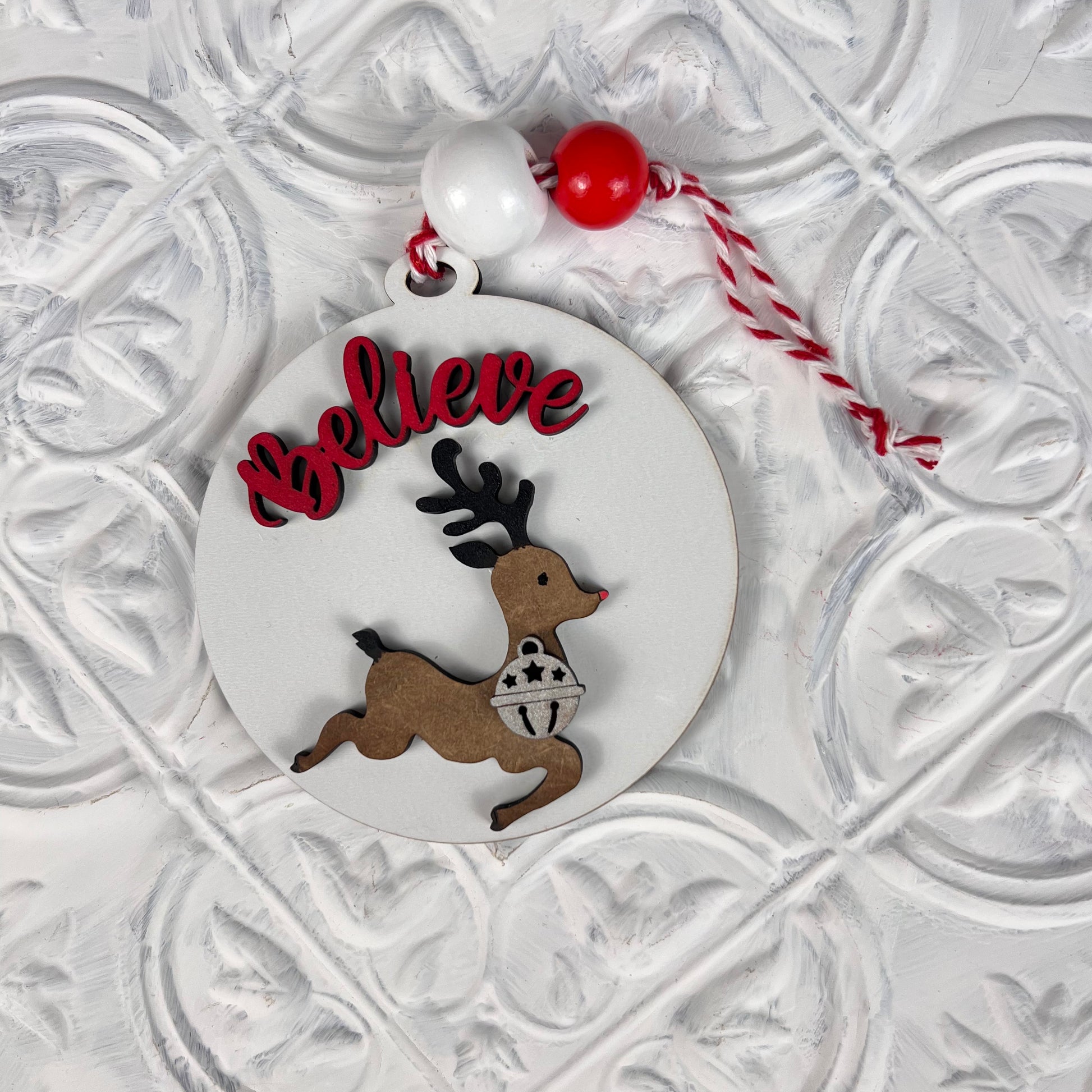 The Round Christmas Ornaments from Janet's Craft Corner feature a cutout of a leaping reindeer in a festive sweater, with the word "Believe" elegantly displayed in red above. Adorned with beads and a red-and-white cord, this intricate decoration adds joy and charm to any DIY Decoration Kit collection.