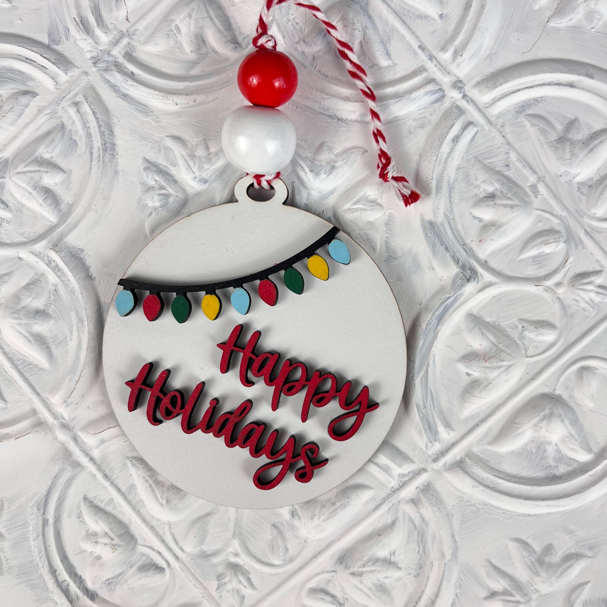 Introducing the Round Christmas Ornaments DIY Decoration Kit from Janet's Craft Corner—featuring a festive ornament with "Happy Holidays" elegantly scripted in red. Accentuated by a string of colorful lights, it hangs beautifully from a coordinating red and white cord with matching beads, enhancing any Christmas display against its textured white background.