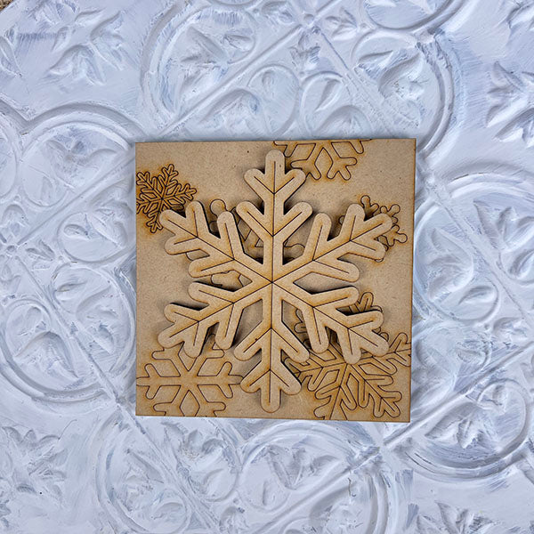 The Winter Interchangeable Squares by Janet's Craft Corner showcases a detailed wooden snowflake cutout elegantly placed on an intricately patterned white textured surface. Ideal for seasonal décor, the snowflake features multiple layers and design variations, emphasizing its intricate craftsmanship and charm in any setting.