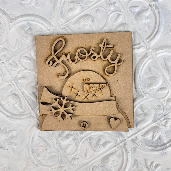 The Winter Interchangeable Squares from Janet's Craft Corner feature a charming wooden cutout of a snowman wearing a top hat and scarf, with the word "Frosty" displayed above. Ideal for tiered trays, this design is embellished with snowflakes, a button, and a heart on a textured white background, perfect for enhancing your seasonal décor.