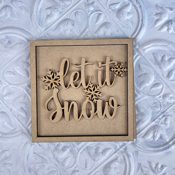 Add a charming touch to your seasonal décor with the Winter Interchangeable Squares from Janet's Craft Corner. This square wooden ornament features the phrase "let it snow" surrounded by snowflakes, making it perfect for winter displays or tiered trays. Its placement on a textured, light-colored background enhances its appeal.