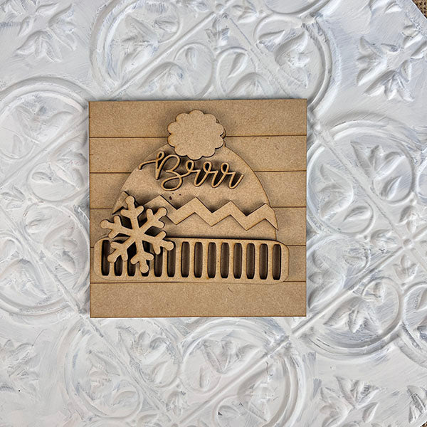 This delightful wooden ornament from Janet's Craft Corner, featuring a winter hat inscribed with "Brrr," is perfect for seasonal décor. It boasts zigzag and stripe patterns, a fluffy pom-pom, and a snowflake design. Ideal for tiered trays or to complement Winter Interchangeable Squares against an intricately patterned white backdrop.