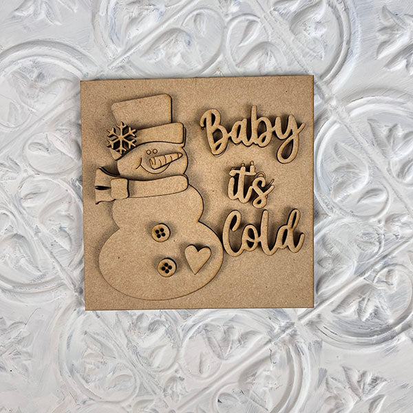 A wooden card intricately carved with a snowman wearing a scarf and top hat, featuring the phrase "Baby it's Cold" in elegant script, is an ideal addition to your seasonal décor. This delightful piece from Janet's Craft Corner fits perfectly into the Winter Interchangeable Squares or on tiered trays, set against its textured white background.