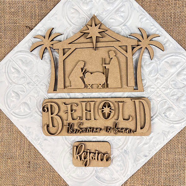 Introducing the Nativity Interchangeable Insert by Janet's Craft Corner, a DIY home decor kit featuring a hand-painted wooden nativity scene with Mary, Joseph, and baby Jesus cradled in a manger framed by palm trees. This versatile decor set includes interchangeable text options: "Behold, the Savior is born" and "Rejoice," displayed on an elegant background to bring warmth and charm to any space.