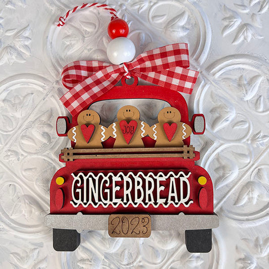 Introducing the Antique Truck Ornament - Painted by Janet's Craft Corner: This enchanting decoration showcases a festive red truck adorned with gingerbread figures in the back and a charming red and white bow. The truck bed features "GINGERBREAD," complemented by a tag marked "2023" against a textured white backdrop, creating the perfect vintage-style charm.