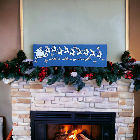 A festive mantel with green garland and red ornaments above a lit fireplace features Janet's Craft Corner’s Santa Sleigh Mantle Shelf Sitter—a blue sign with white reindeer and sleigh reading "and to all a goodnight.