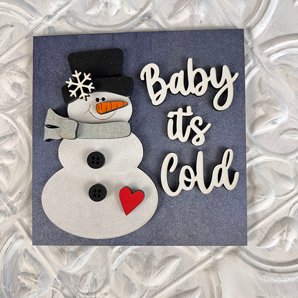 Introducing the Winter Interchangeable Squares by Janet's Craft Corner: a festive greeting card that's perfect for seasonal décor. It features a charming snowman crafted from paper cutouts, complete with a black hat and gray scarf. Adorned with buttons and a heart on its body, along with a snowflake design on its face, this card captures the spirit of winter. The text "Baby it's Cold" is elegantly written in white cursive letters, making it an ideal choice for your holiday decorations.